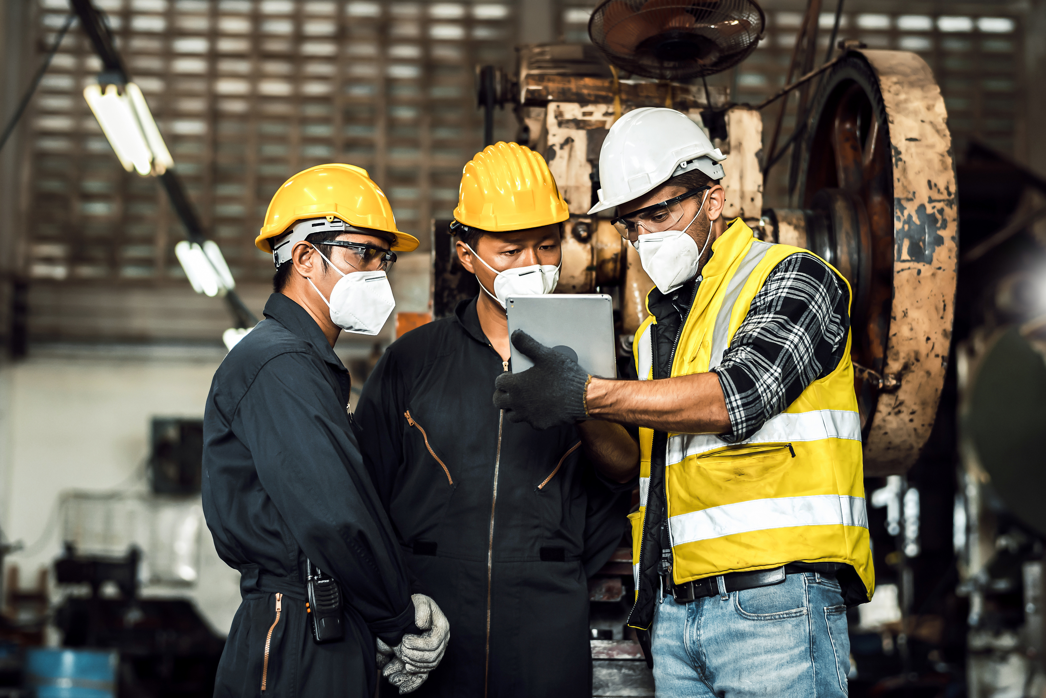 Three employees in jumpsuits and safety gear examining safety protocols on a tablet.