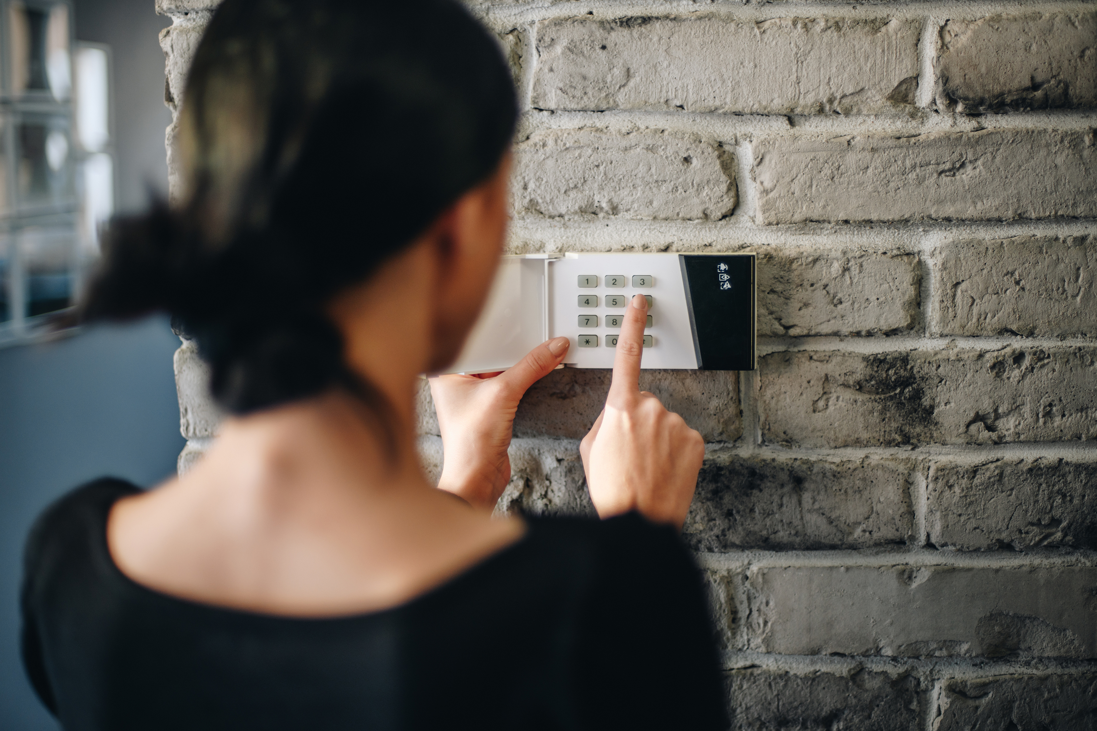 Over-the-shoulder view of woman entering code into wall-mounted security system panel.