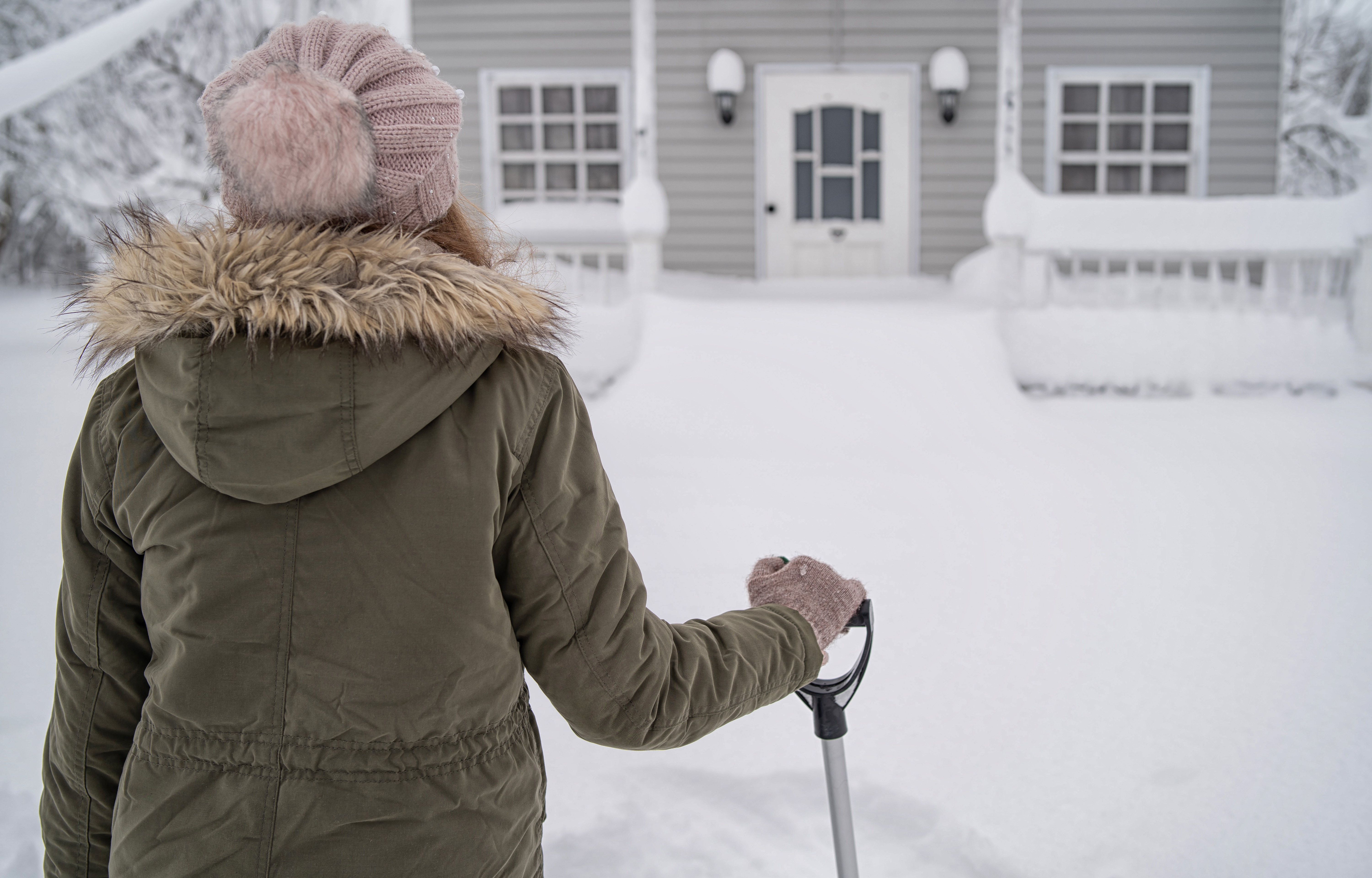 Woman in winter hat and coat looking out over snowy scene in front of house.