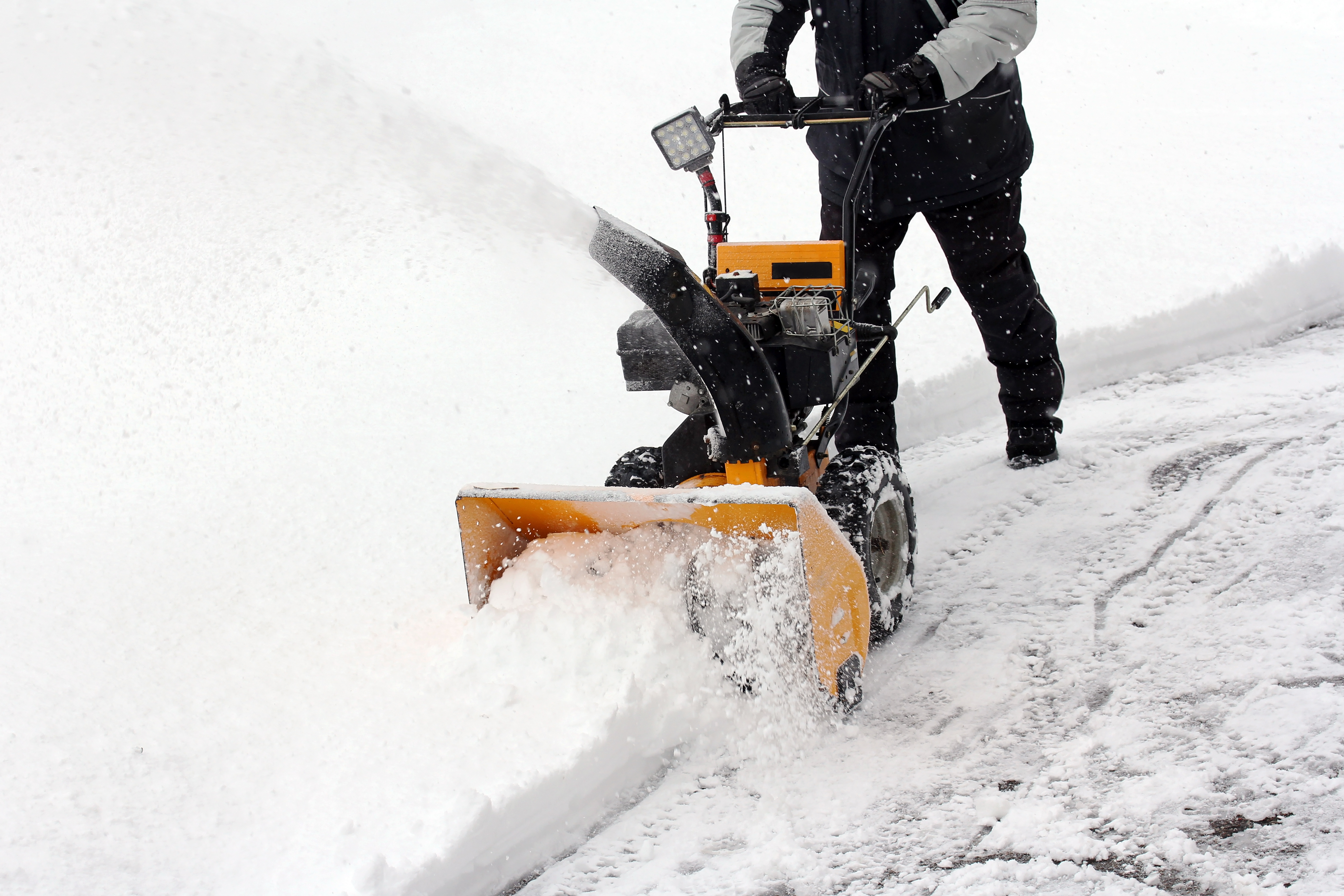 A person bundled in winter coat and gloves using a snowblower to remove snow from a driveway.