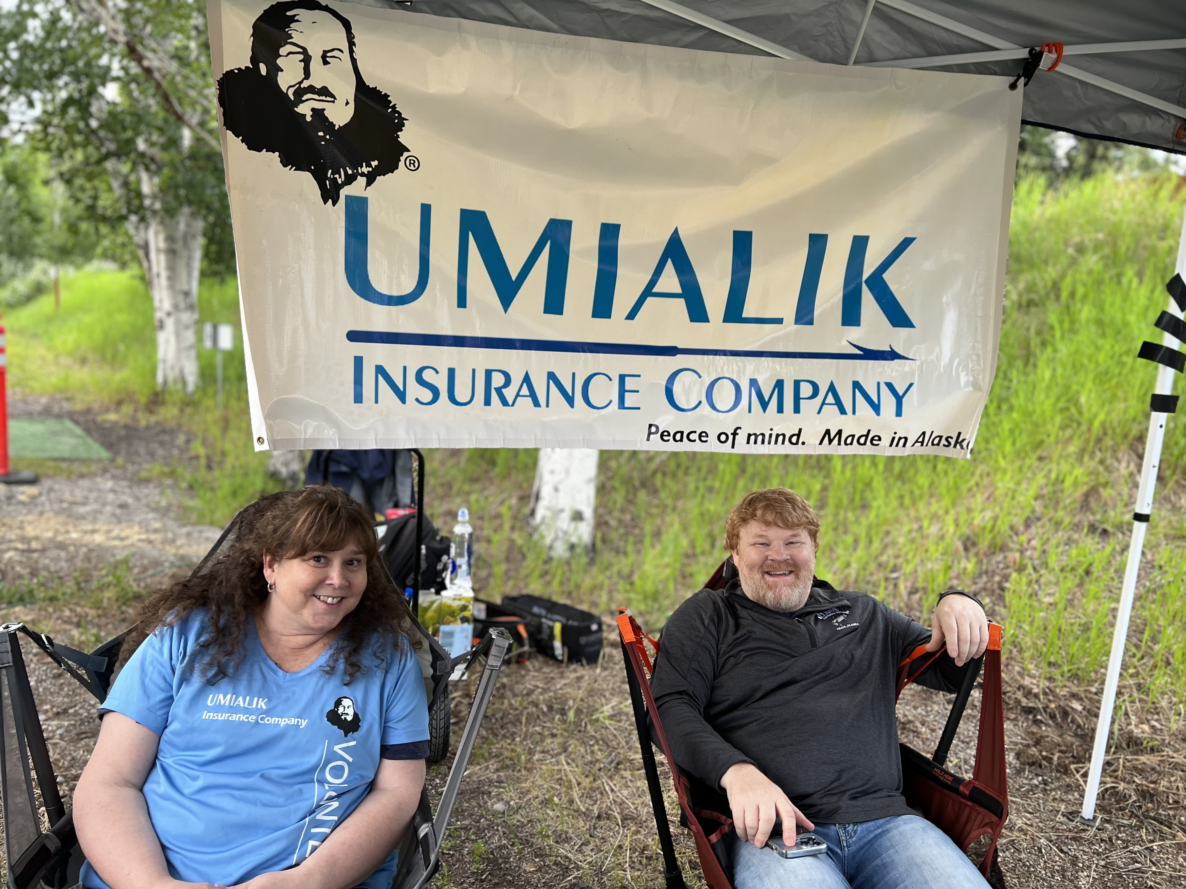 Two Umialik employees sitting outdoors in camp chairs with a large Umialik-branded banner hanging behind them.