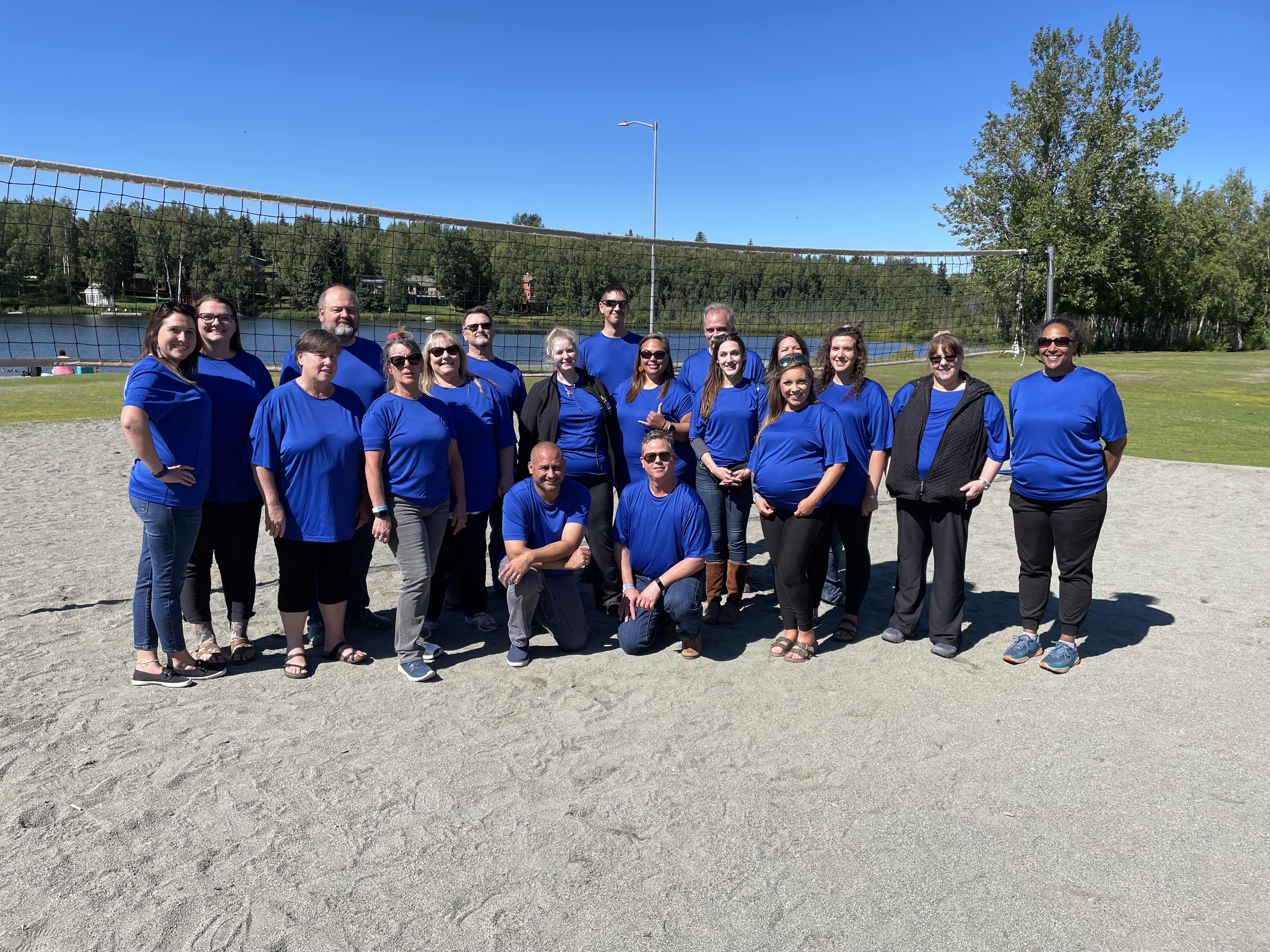 Group of Umialik employees in matching blue shirts posing together outdoors on a sand volleyball court.